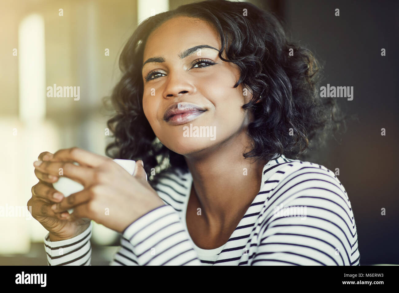 Young African woman sitting alone at a table in a cafe enjoying a fresh cup of coffee and looking deep in thought Stock Photo