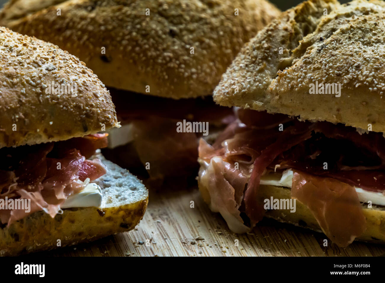 Three serrano ham sandwiches with wholegrain bread and brie cheese laying on a wooden board with small grain and bread crumbs Stock Photo