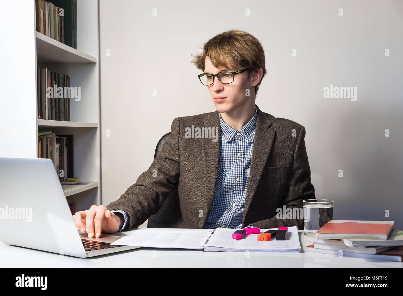 Handsome young person in reading eyeglasses at modern minimalistic workplace. Young male student works with books and laptop on home assignment or pro Stock Photo