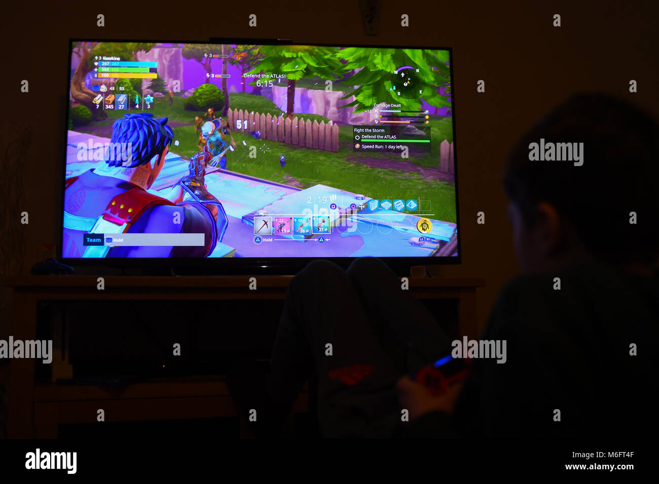 A young boy plays the computer game Fortnite on a PS4 in a darkened room on a large TV. Stock Photo