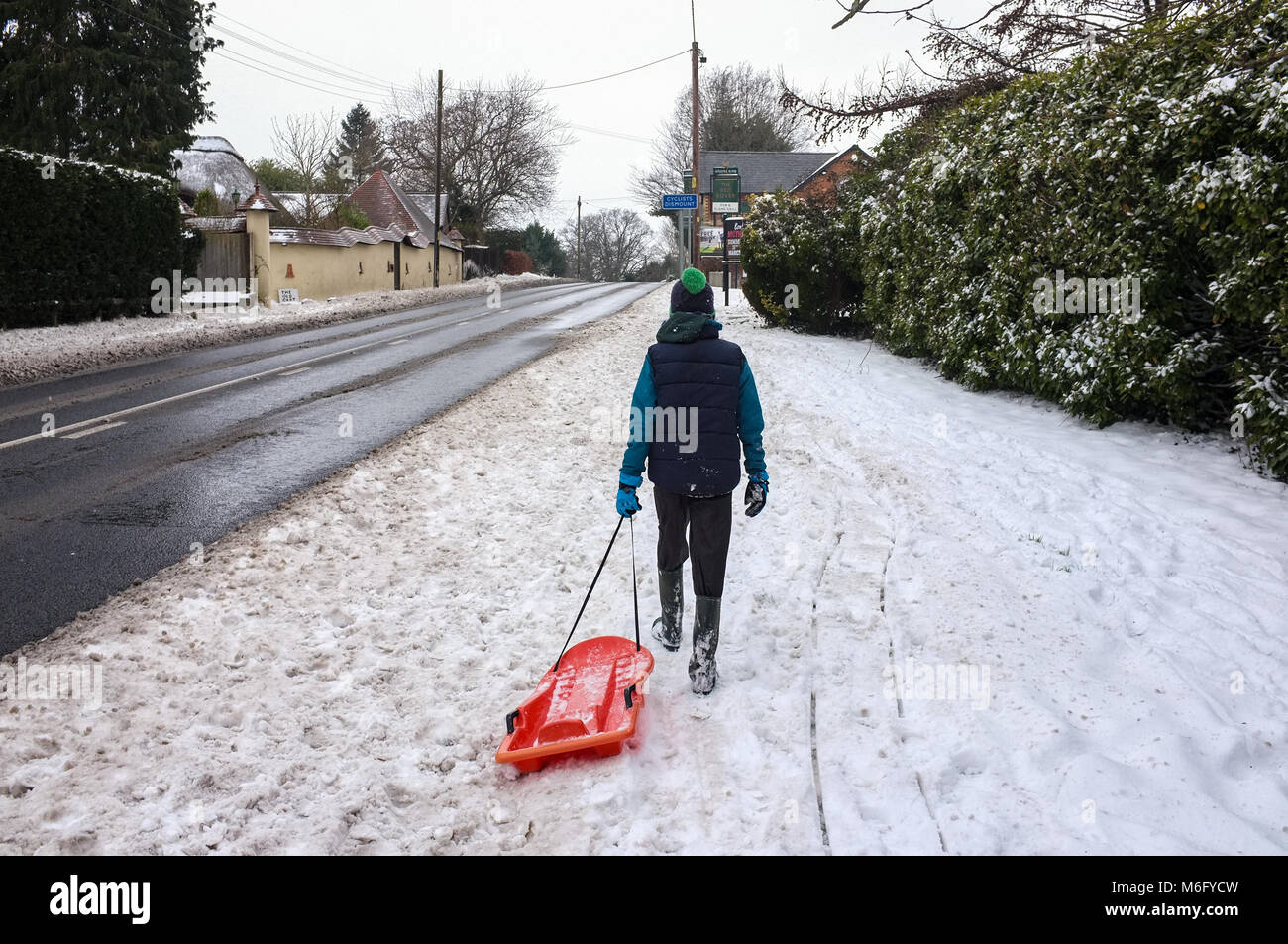 A boy walks on a journey alone on the snow covered path pulling a sledge alongside the A36 towards the Red Rover public house West Wellow England. Stock Photo