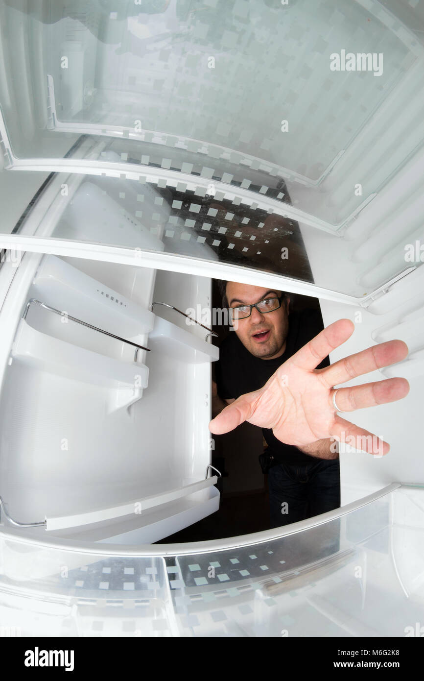 Funny scene of hungry young man opening the door of the fridge and reaching for food, view from inside the freezer Stock Photo