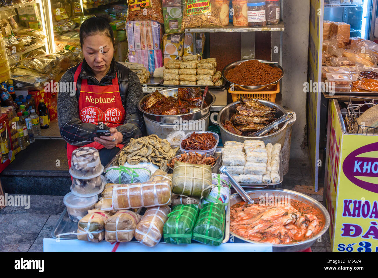 A woman looks at her Smartphone while waiting for customers to her street food stall on a footpath in Hanoi, Vietnam Stock Photo