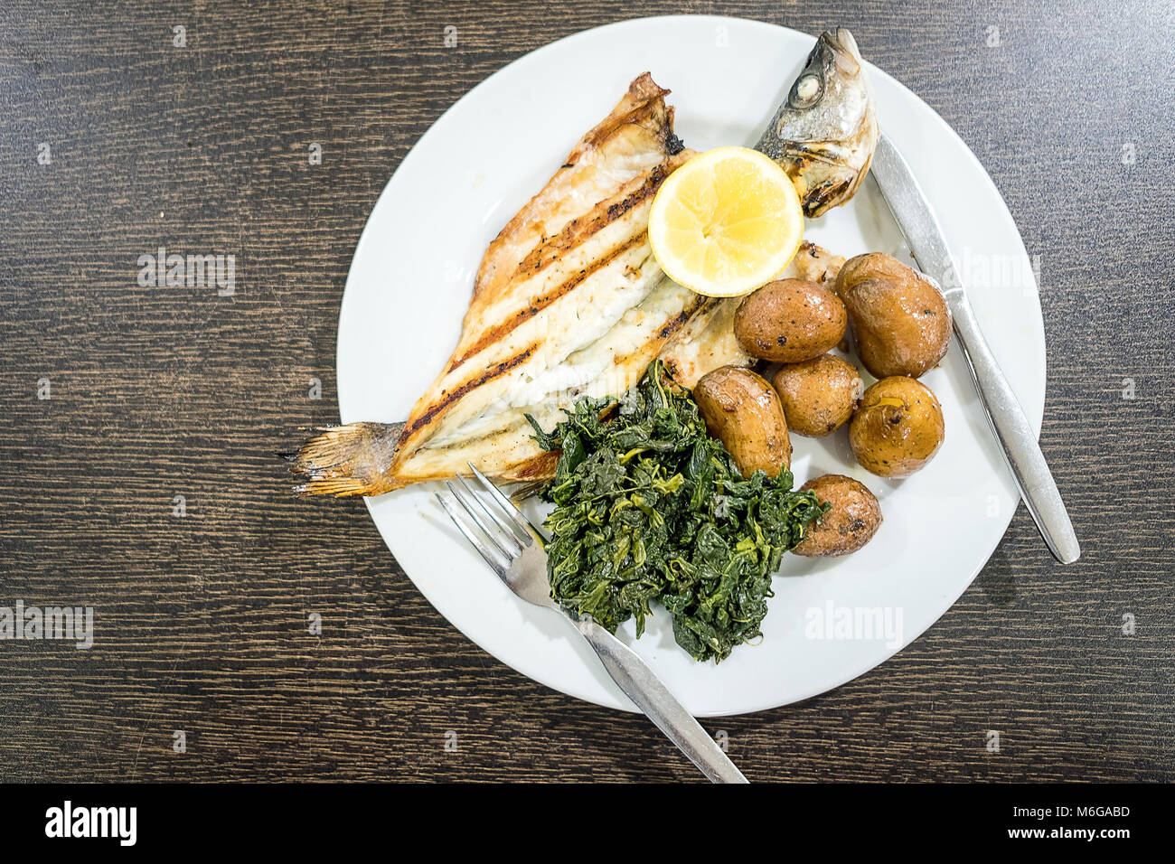 Barbecued sea bass served with lemon, baked potatoes and spinach on white plate Stock Photo