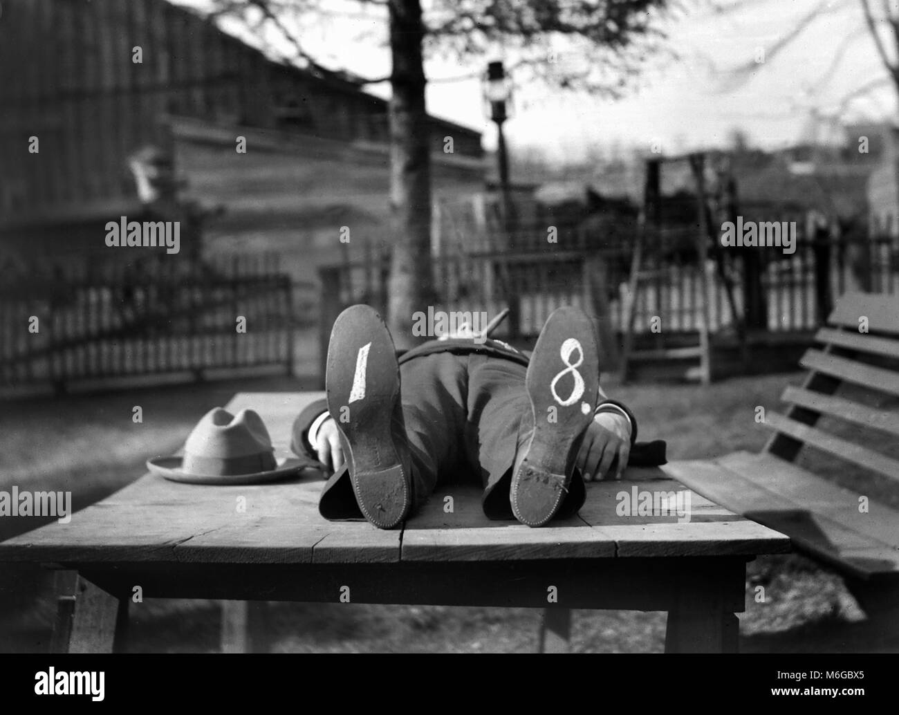 A hatless man rests on an outdoor table with cigar in mouth and writing on his shoes, ca. 1910. Stock Photo