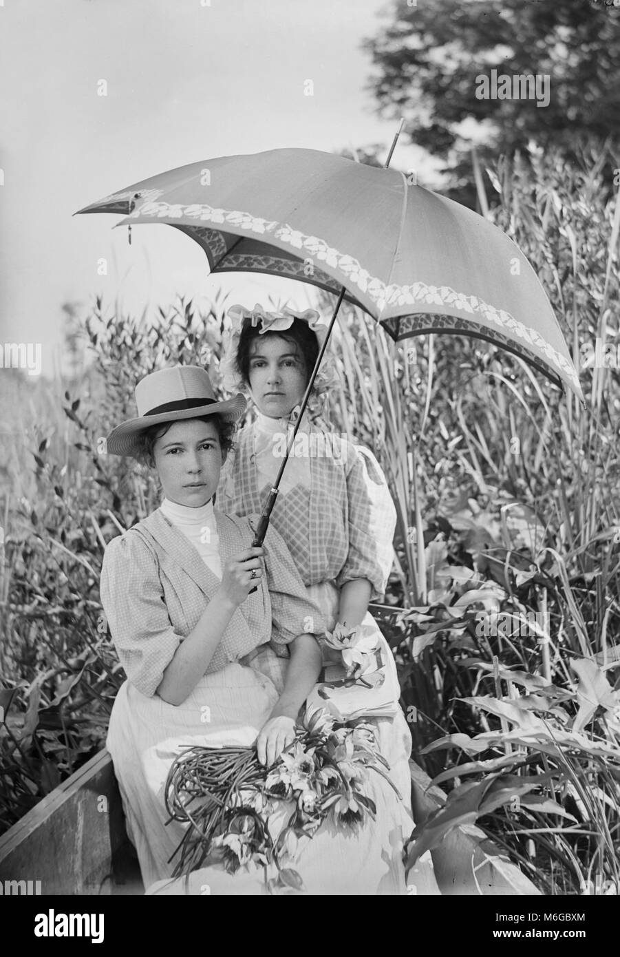 Sisters strike a rather Victorian-looking pose together in the bow of a boat under a parasol, ca. 1900. Stock Photo