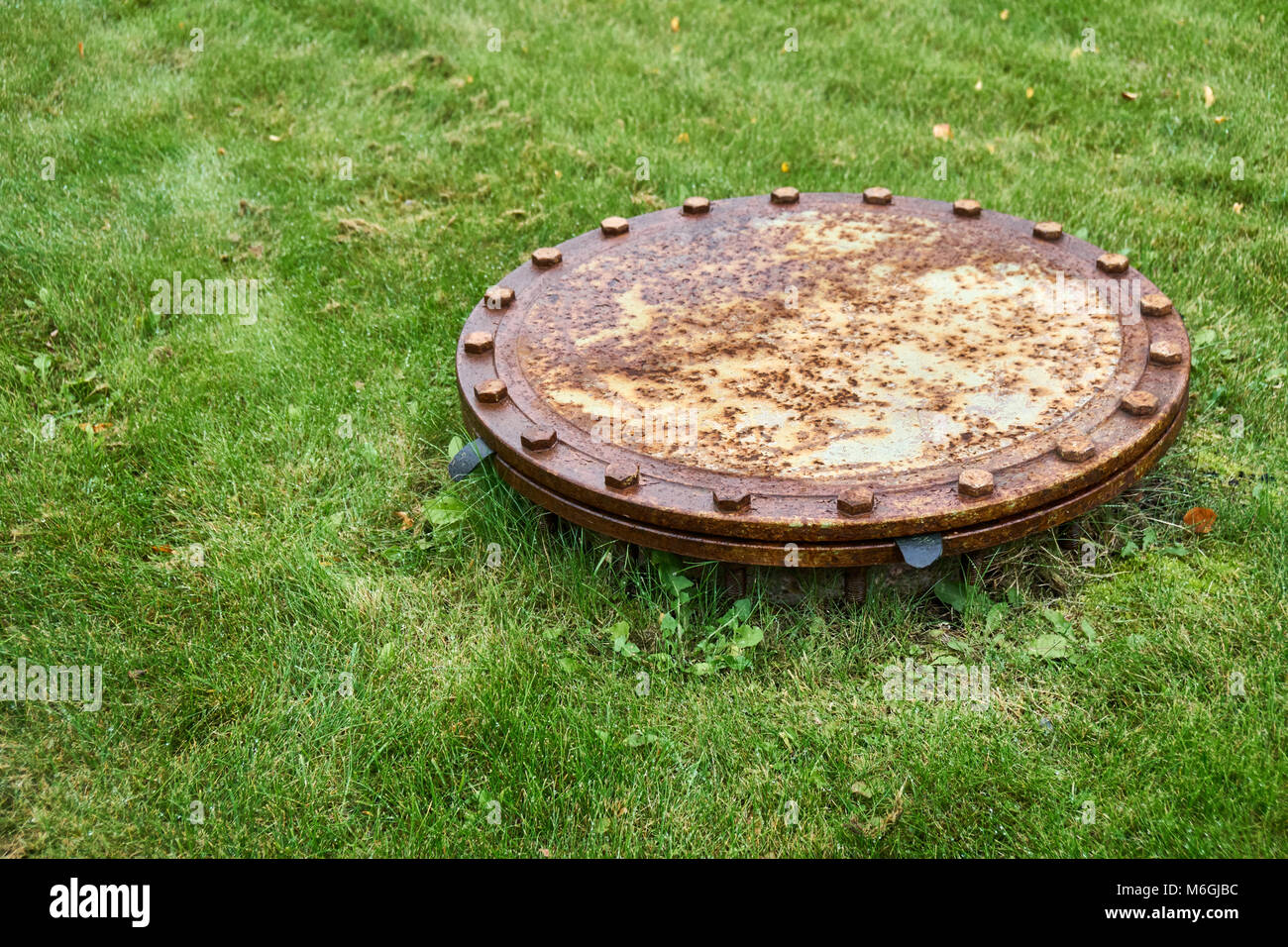 Outdated round manhole rusty metal cover on rain drainage system located on lawn with green lush grass closeup Stock Photo