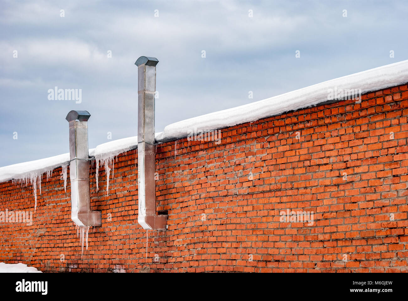 Red brick wall with ventilation pipes atop a concrete fence adorned with icicles, hinting at cold weather conditions Stock Photo