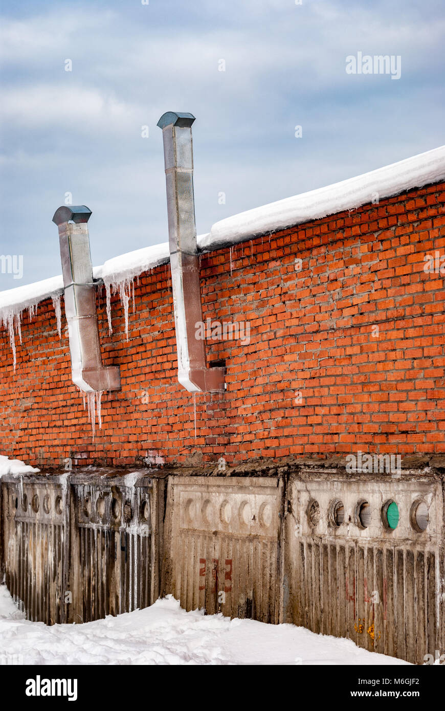 Red brick wall with ventilation pipes atop a concrete fence adorned with icicles, hinting at cold weather conditions Stock Photo