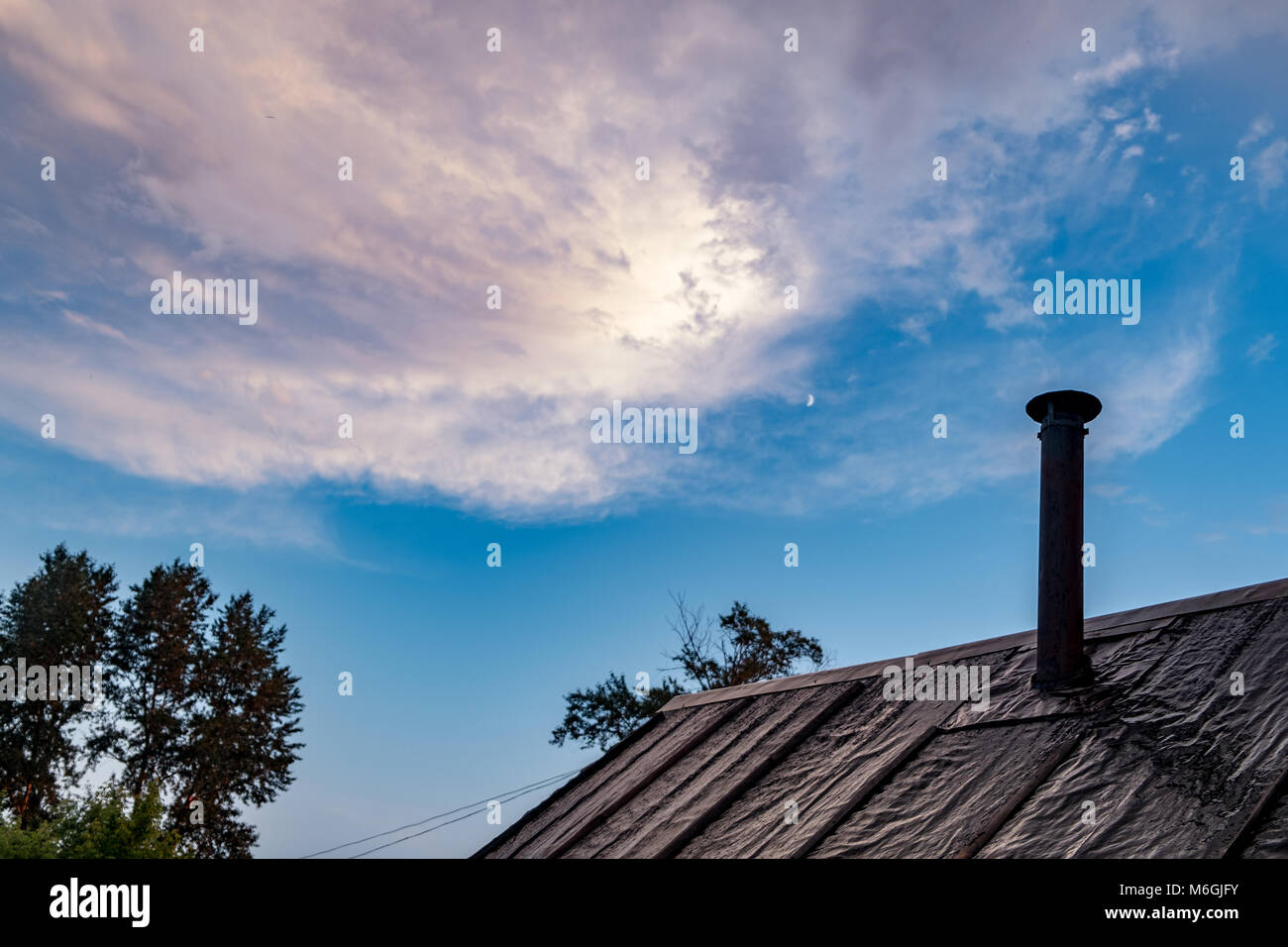 Early evening sky with mix of pink and blue hues. In foreground worn rooftop with roofing felt and metal chimney contrasts with natural backdrop Stock Photo