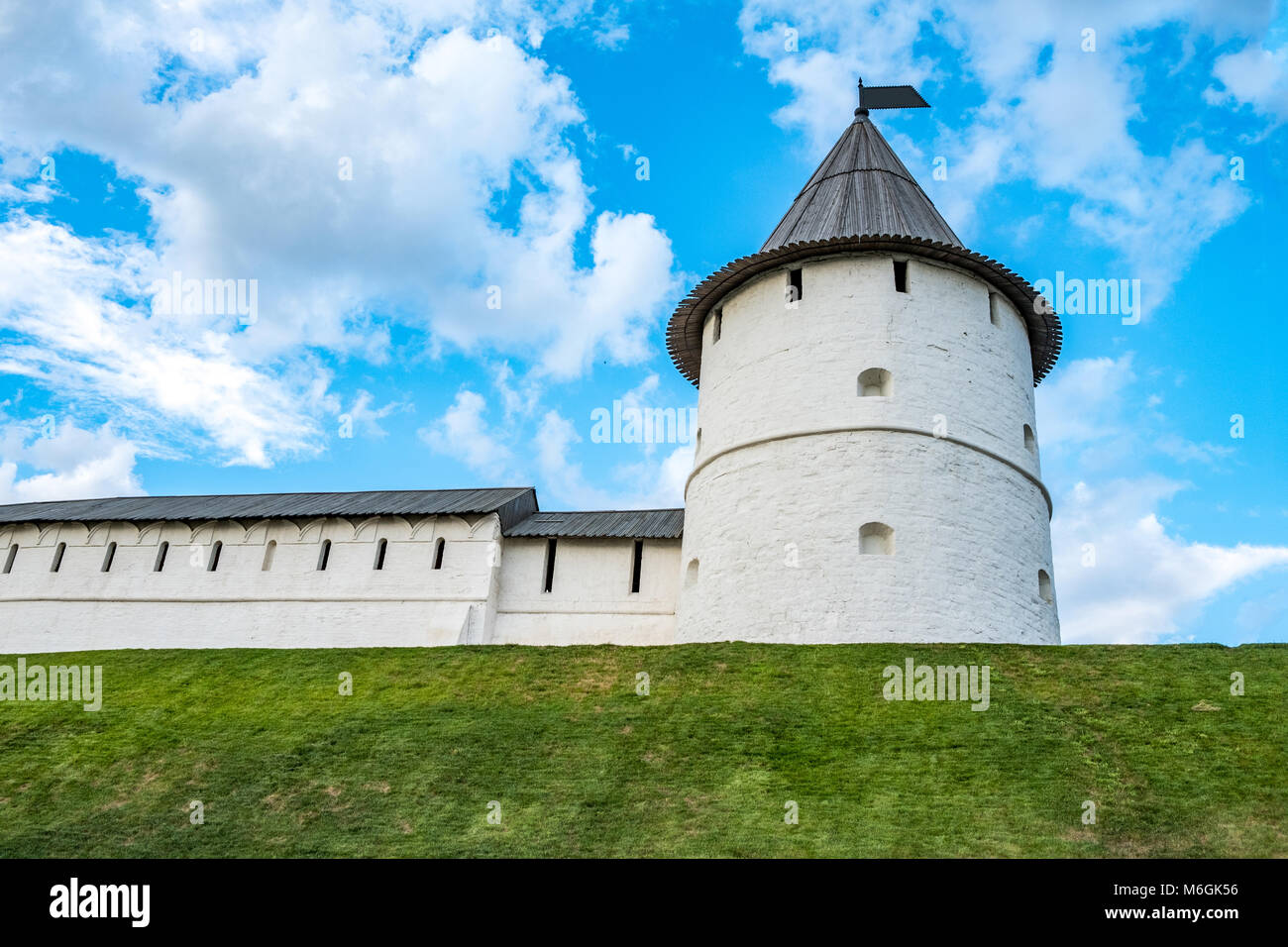 Majestic white medieval fortress tower with a conical roof atop a grassy hill, symbolizing historical protection under a vast blue sky with clouds Stock Photo