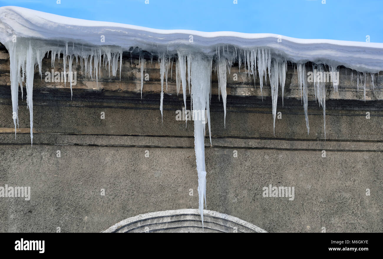 Spring icicles hanging down from snow covered roof on a blue sky background. Icicles are very dangerous for people's life/ dangerous seasonal weather Stock Photo