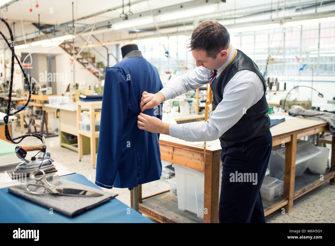 Tailor working on a partially completed new blue jacket on a mannequin in his large spacious workshop tucking in the pleats at the back with his hands Stock Photo