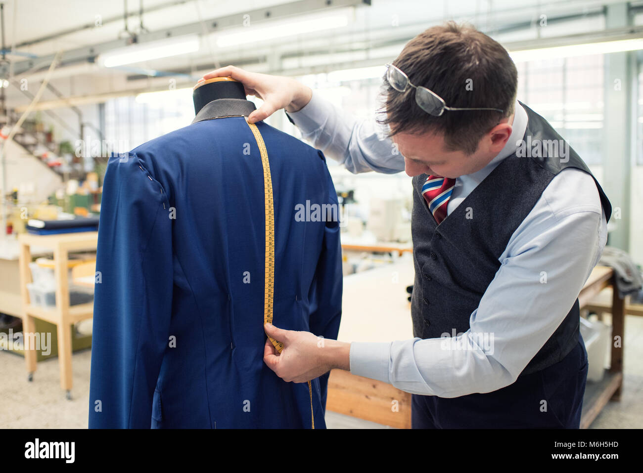 Tailor measuring the length of the back of a new blue jacket on a mannequin that he is busy sewing in his workshop using a tape measure Stock Photo