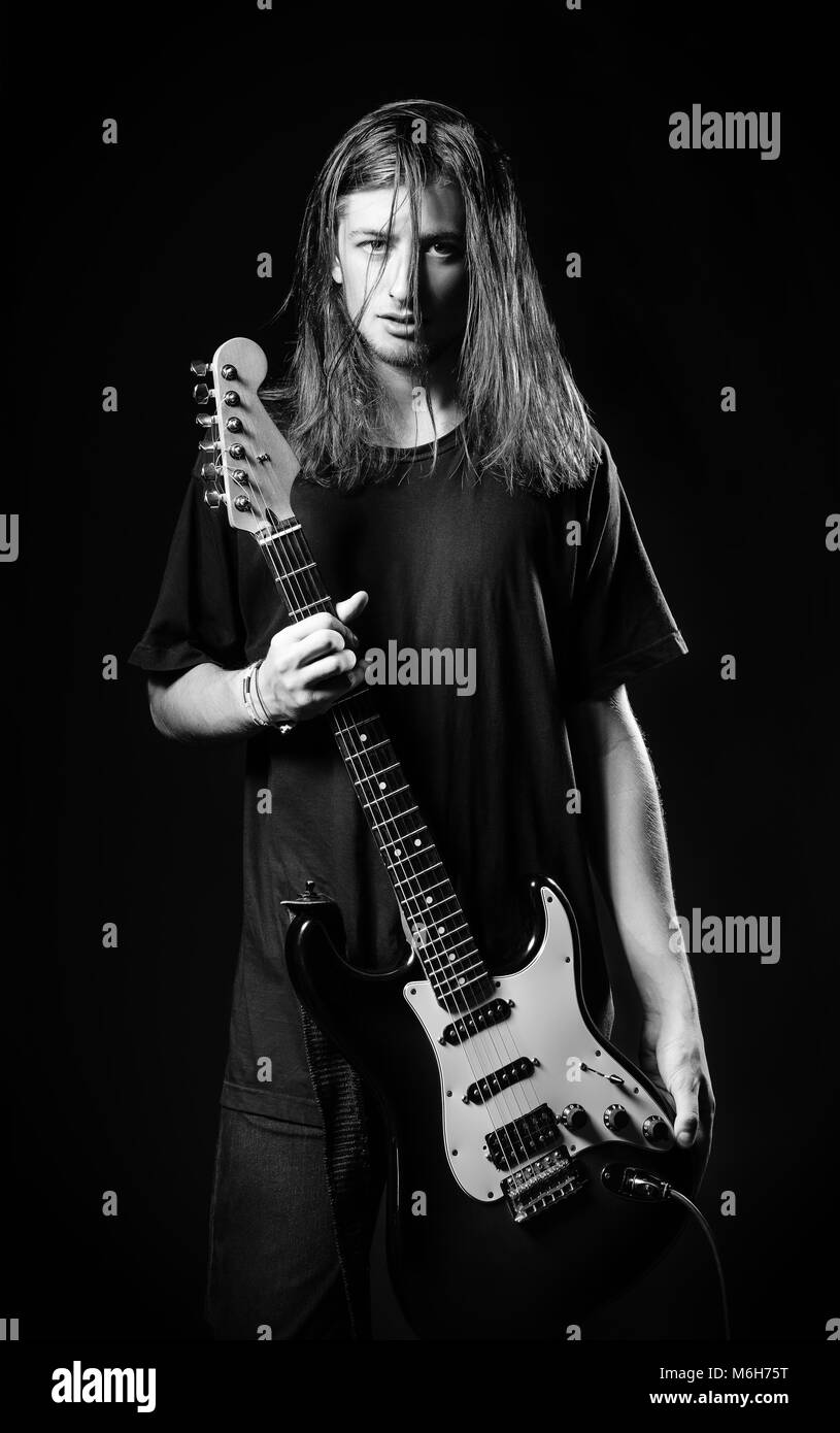 Dramatic studio portrait: a handsome long-haired young man (rock musician) holds electric guitar in hands. Black and white Stock Photo