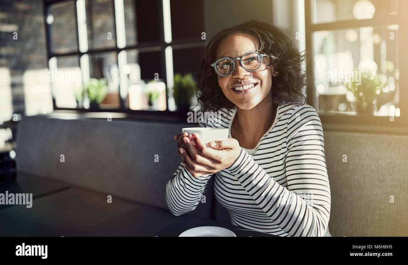 Smiling young African woman sitting at a table in a cafe drinking a cup ...