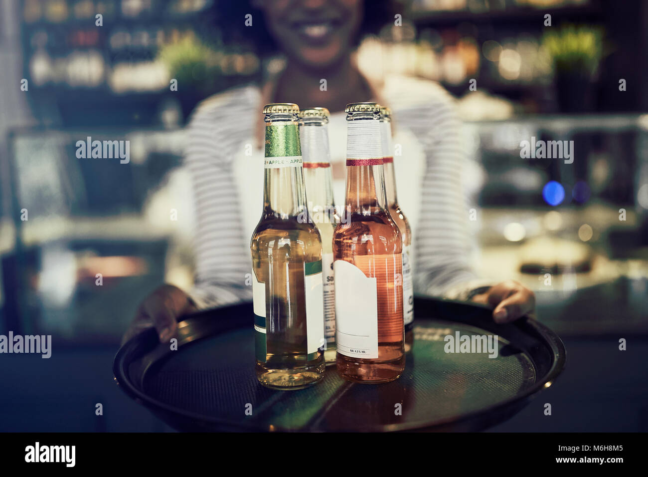Smiling young African server standing in a trendy restaurant holding a tray of bottled drinks Stock Photo