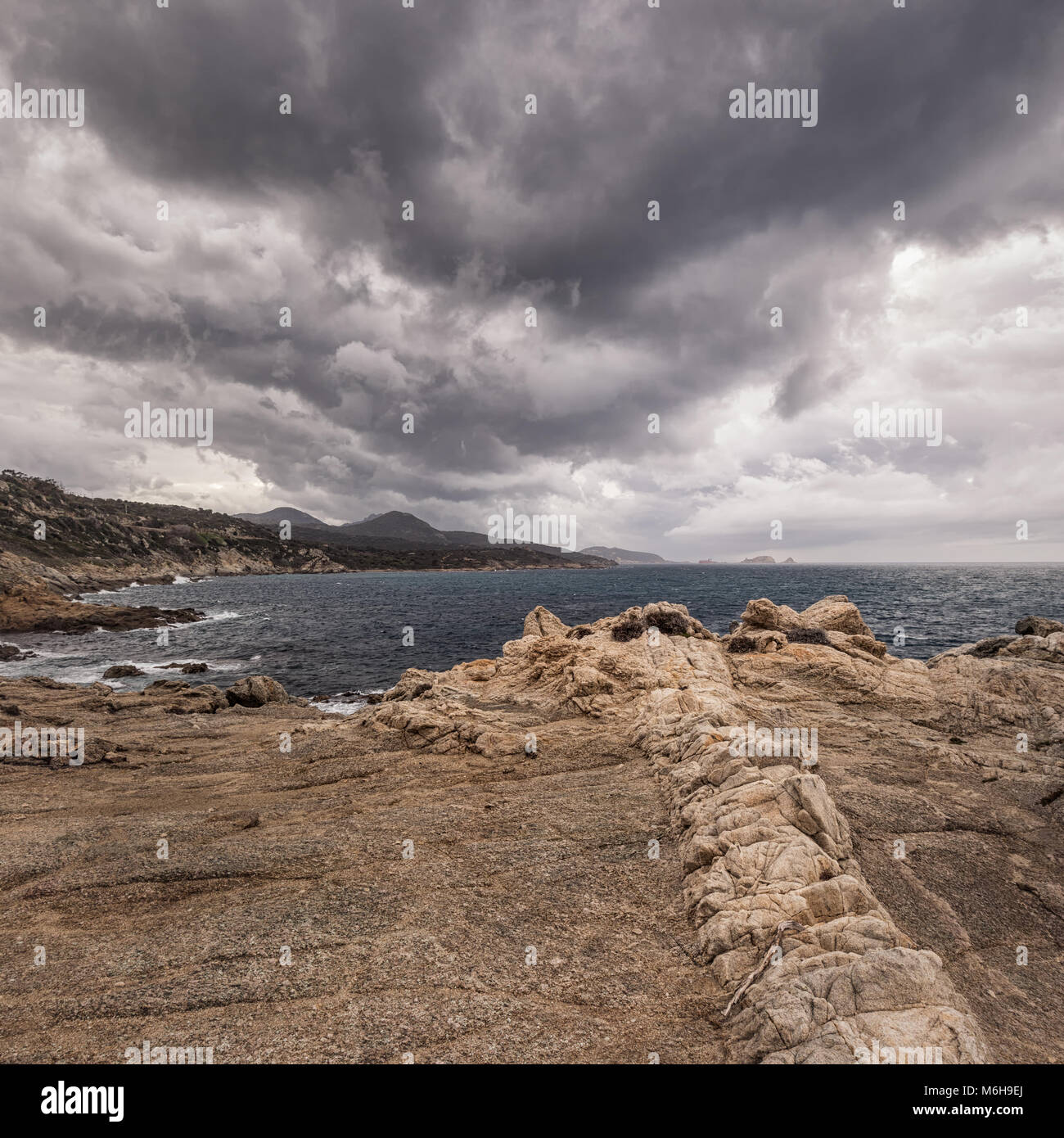 A line of rock striations on the coast of Corsica with Ile Rousse in the distance under dark storm clouds Stock Photo