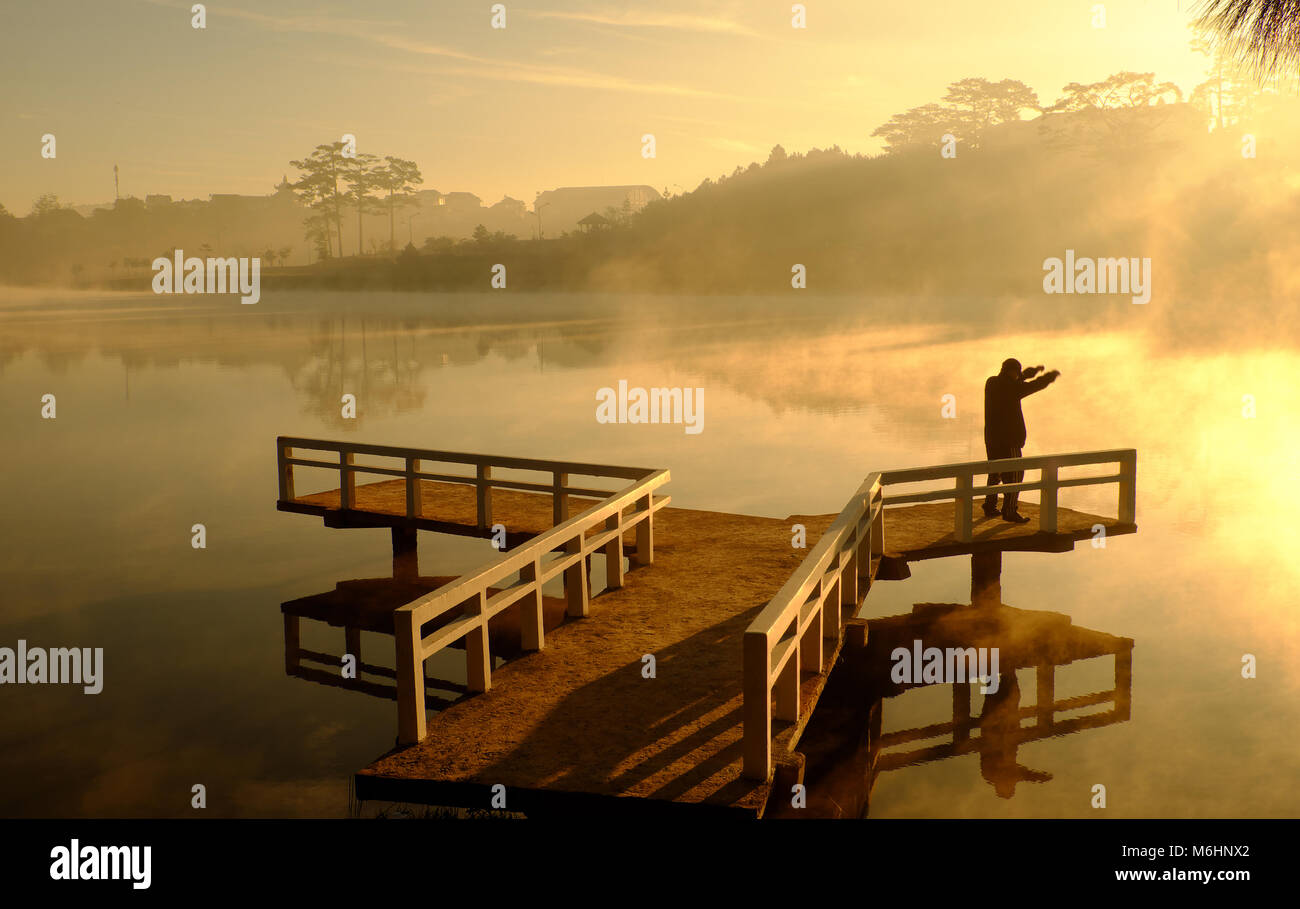 Amazing landscape of Da Lat city at sunrise, silhouette of man do exercise on small bridge reflect on surface water of lake, fog evaporate from pond Stock Photo