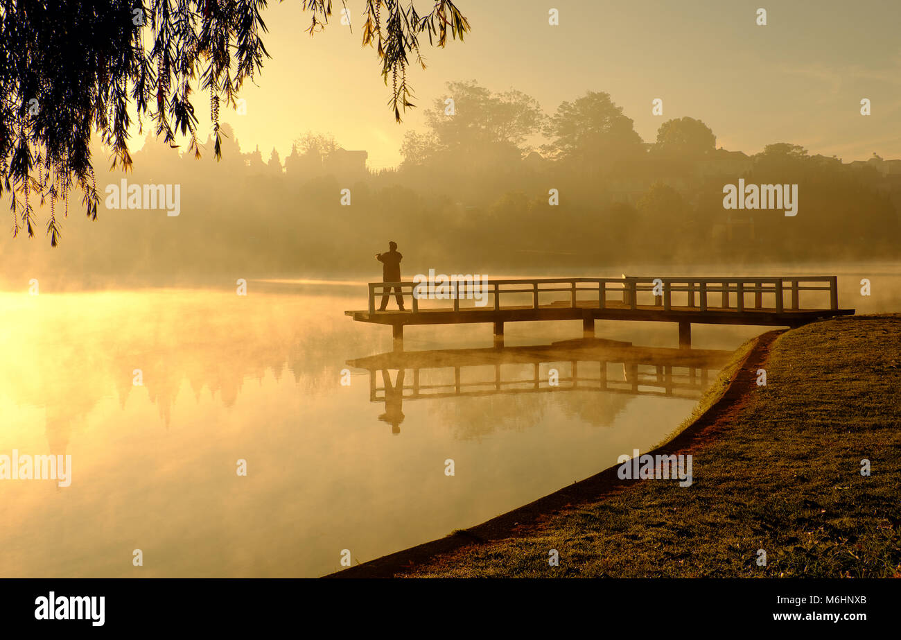 Amazing landscape of Da Lat city at sunrise, silhouette of man do exercise on small bridge reflect on surface water of lake, fog evaporate from pond Stock Photo