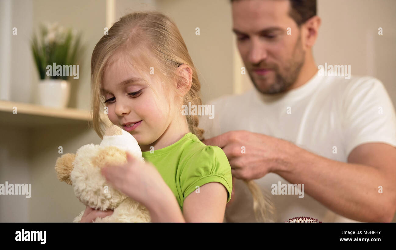 Happy daughter waiting father braiding her blonde hair, playing with teddy-bear Stock Photo