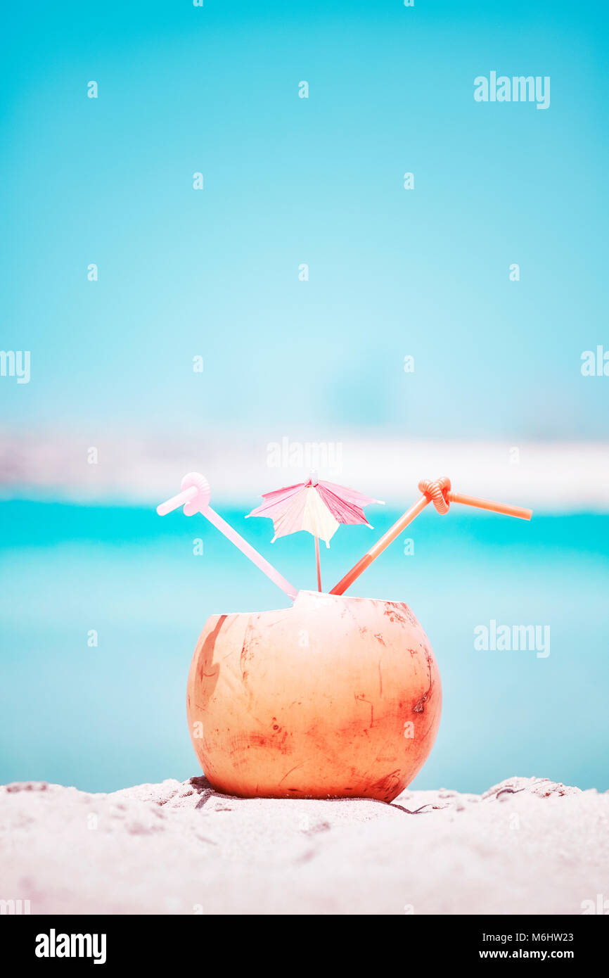 Retro toned picture of a coconut with two straws and colorful umbrella on a beach, summer holiday concept, selective focus. Stock Photo