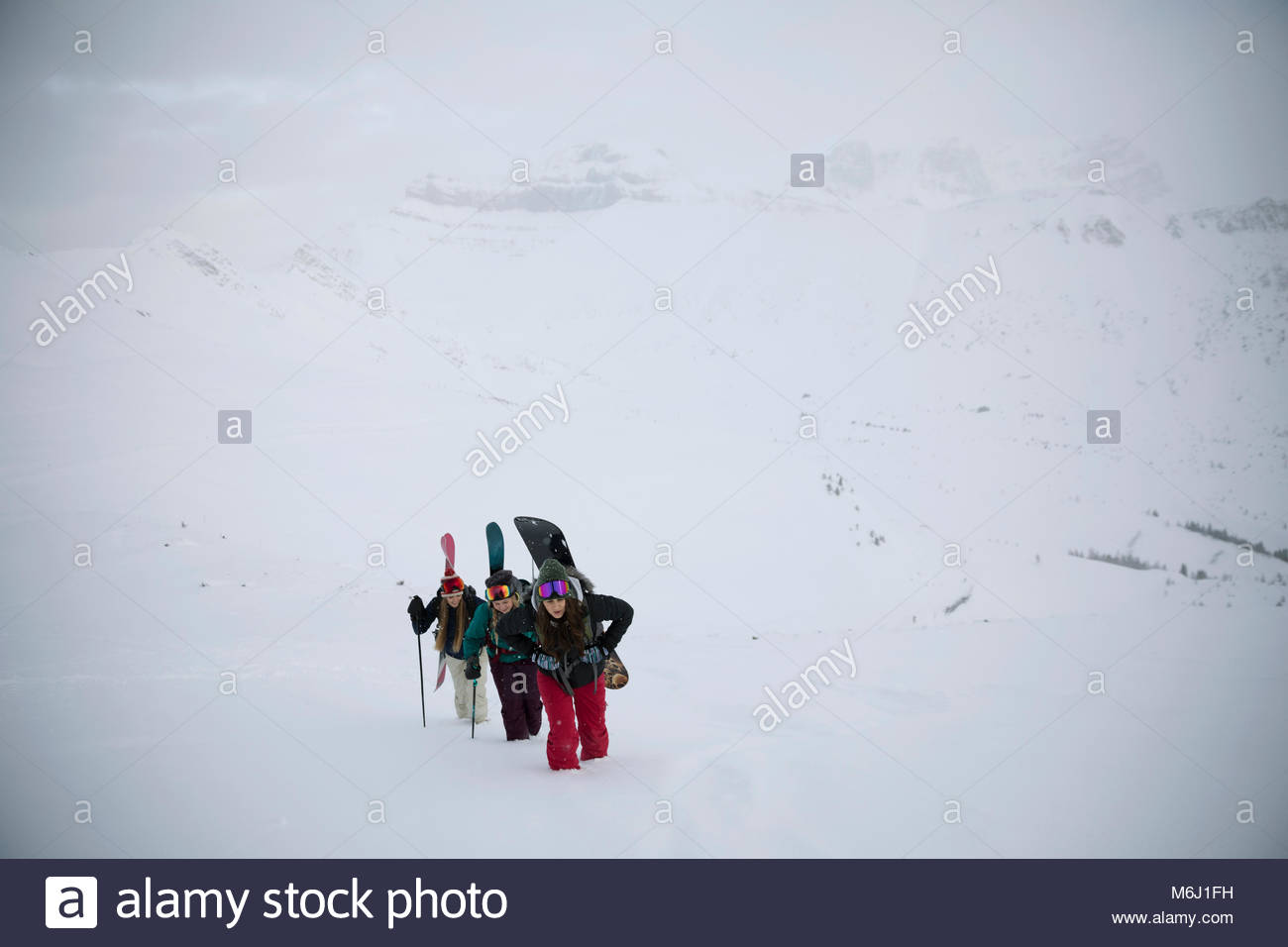 Female skier and snowboarder friends hiking up snow covered hill Stock Photo