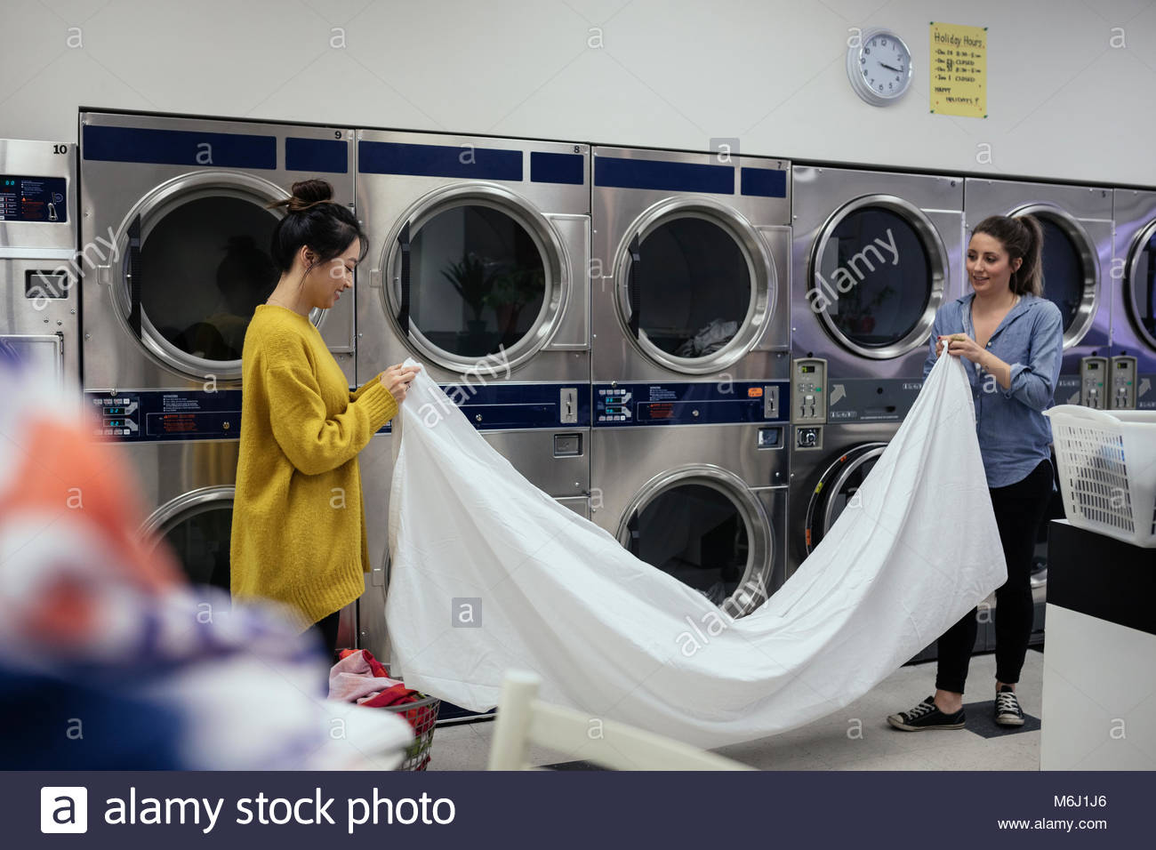 Young women friends folding sheet, doing laundry at laundromat Stock