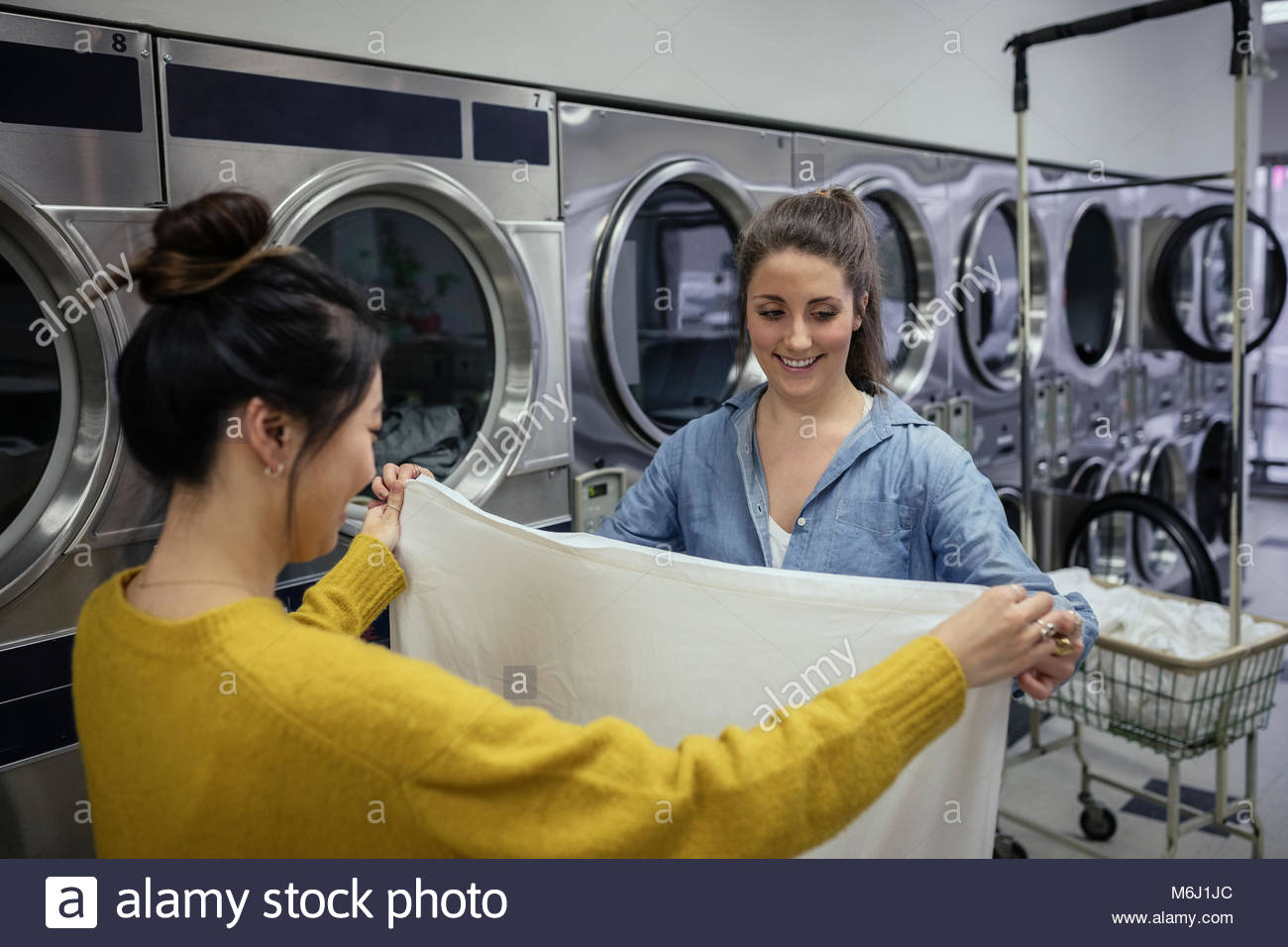 Young women friends folding sheet, doing laundry at laundromat Stock