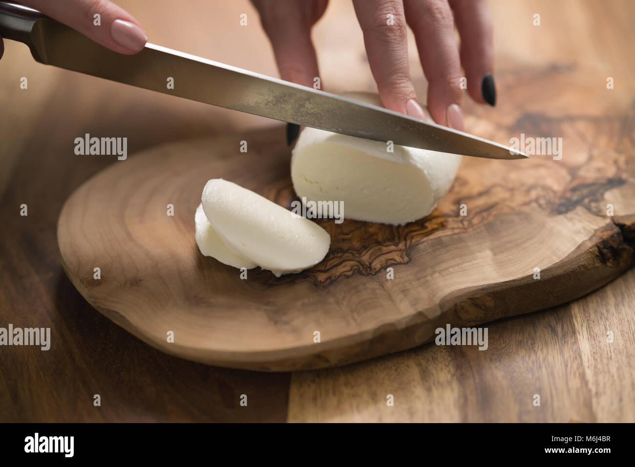 female teen hand slicing mozzarella cheese with knife on wooden board