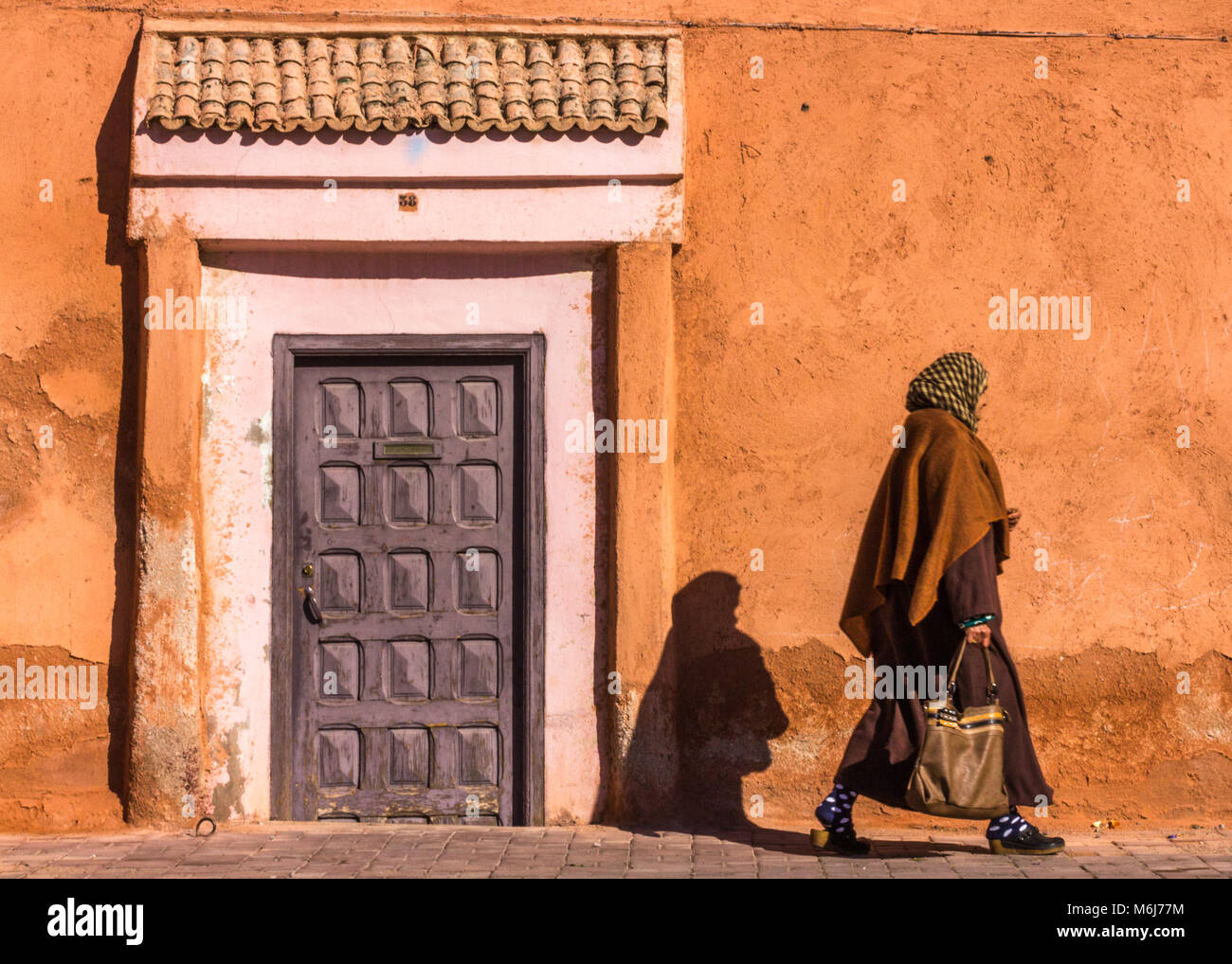 A local woman walks past a low doorway set into ancient walls of the Medina, Marrakesh. Her shadow is cast on the wall Stock Photo