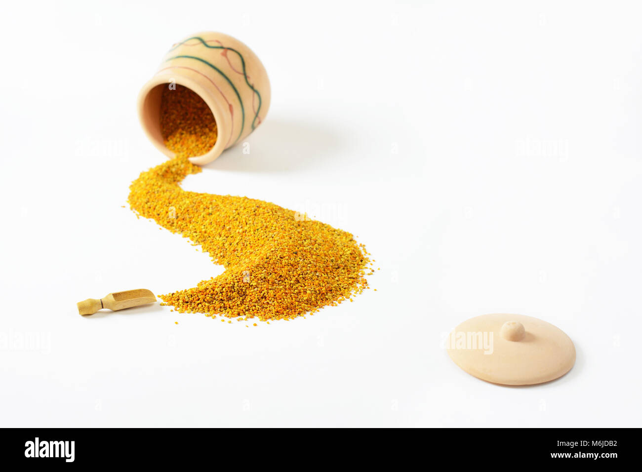 Bee pollen poured out from a small glazed clay pot is making a trail that leads to one little wooden scoop. In the foreground lies a lid. Stock Photo