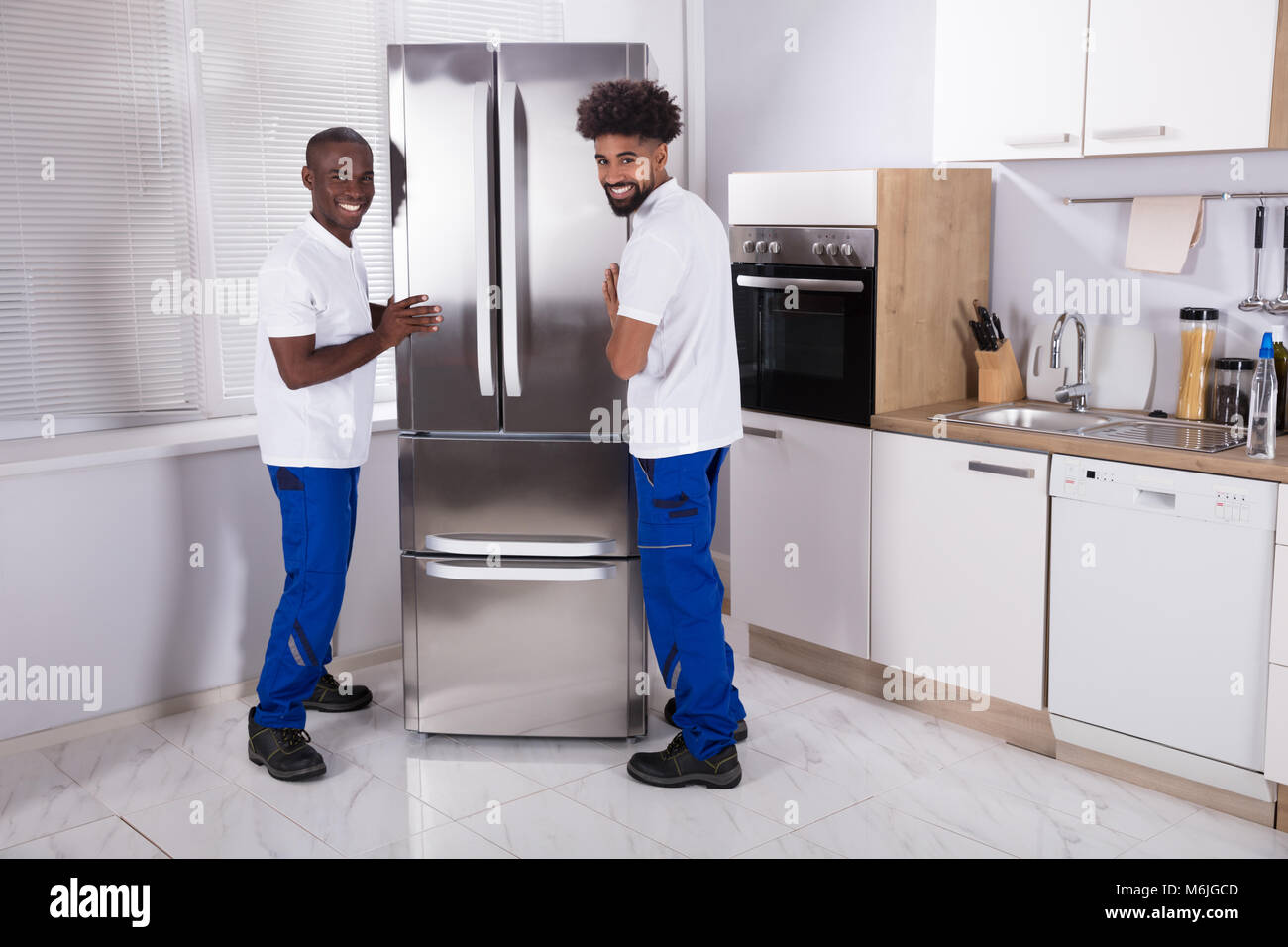 Two Male Movers In Uniform Fixing The Freezer In The Kitchen At Home Stock Photo