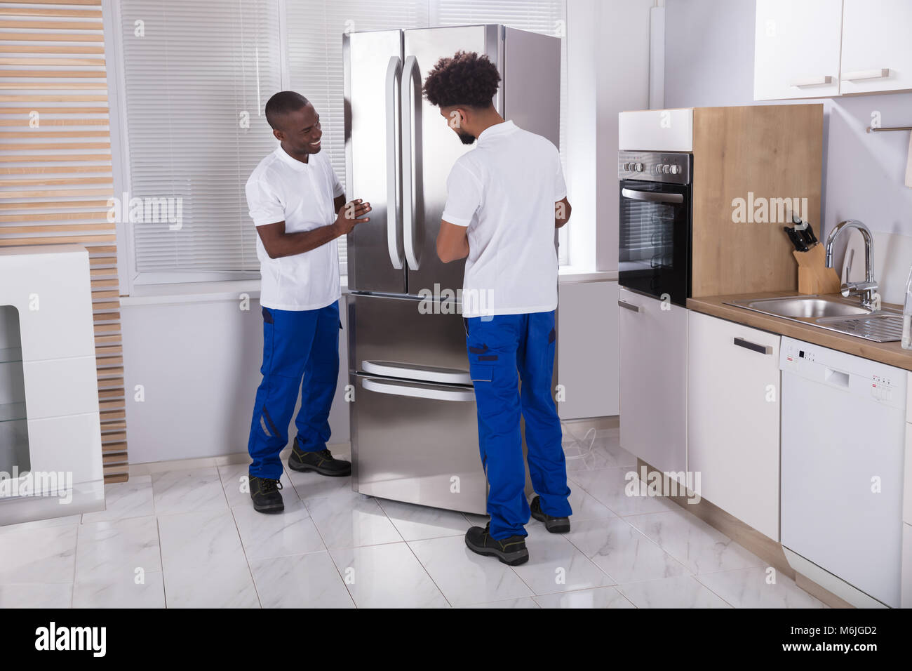 Two Male Movers In Uniform Fixing The Freezer In The Kitchen At Home Stock Photo