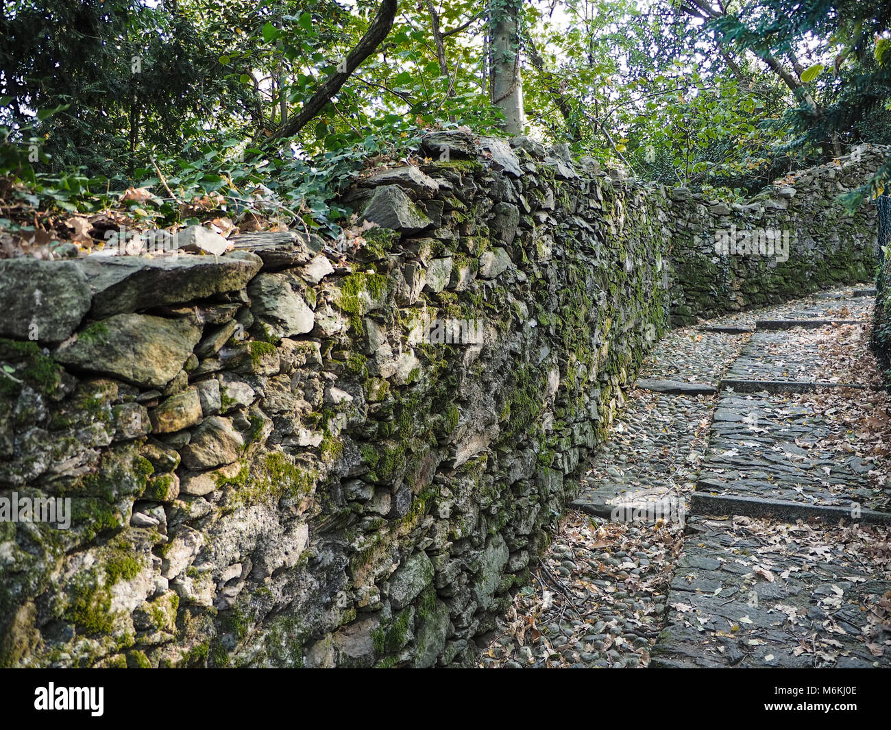 path uphill in cobblestone that runs along a stone wall and a forest Stock Photo