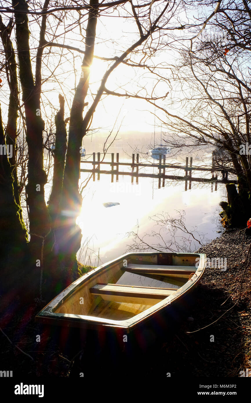 lake Windermere, Lake District, UK, a rowing boat on the shoreline with a wooden jetty, early morning view across the flat calm lake forming a complet Stock Photo