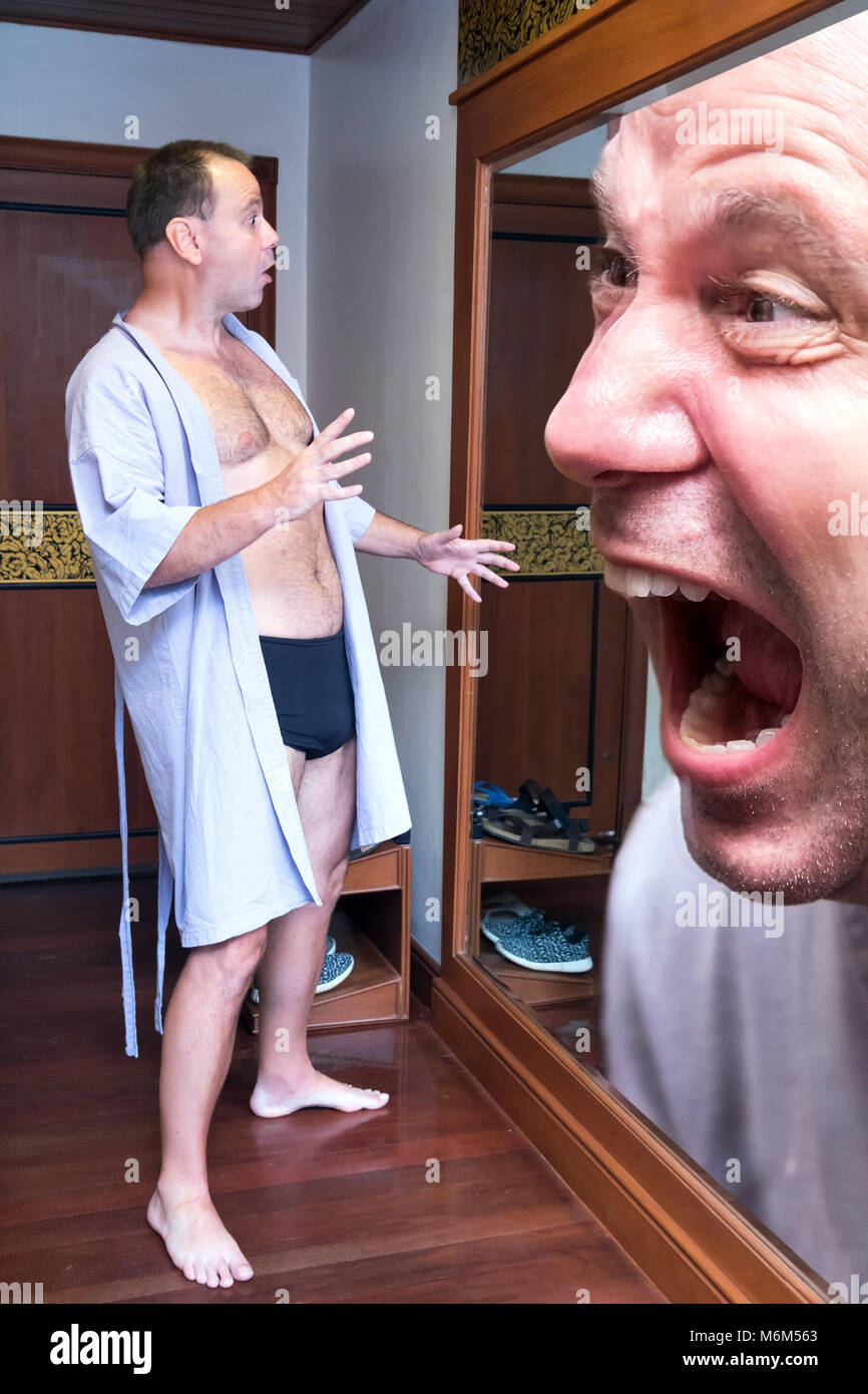 The shocked frightened man in a bathrobe looks at own large screaming head in the mirror. Reflection of man in the mirror screaming at himself. Stock Photo