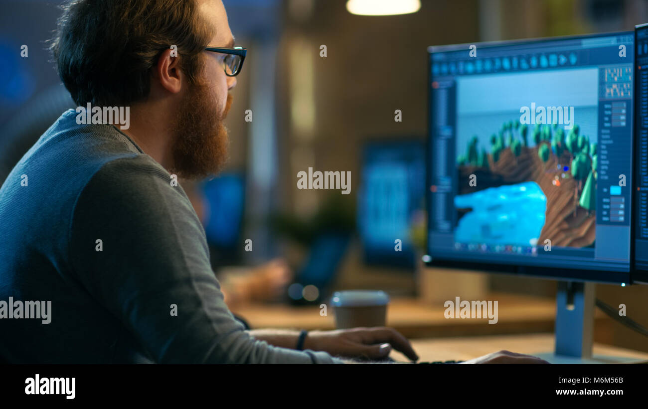 Male Game Developer Works on a New Level Design. He's Handsome Bearded Man with Glasses and Works in a Creative Office with other Talented People. Stock Photo