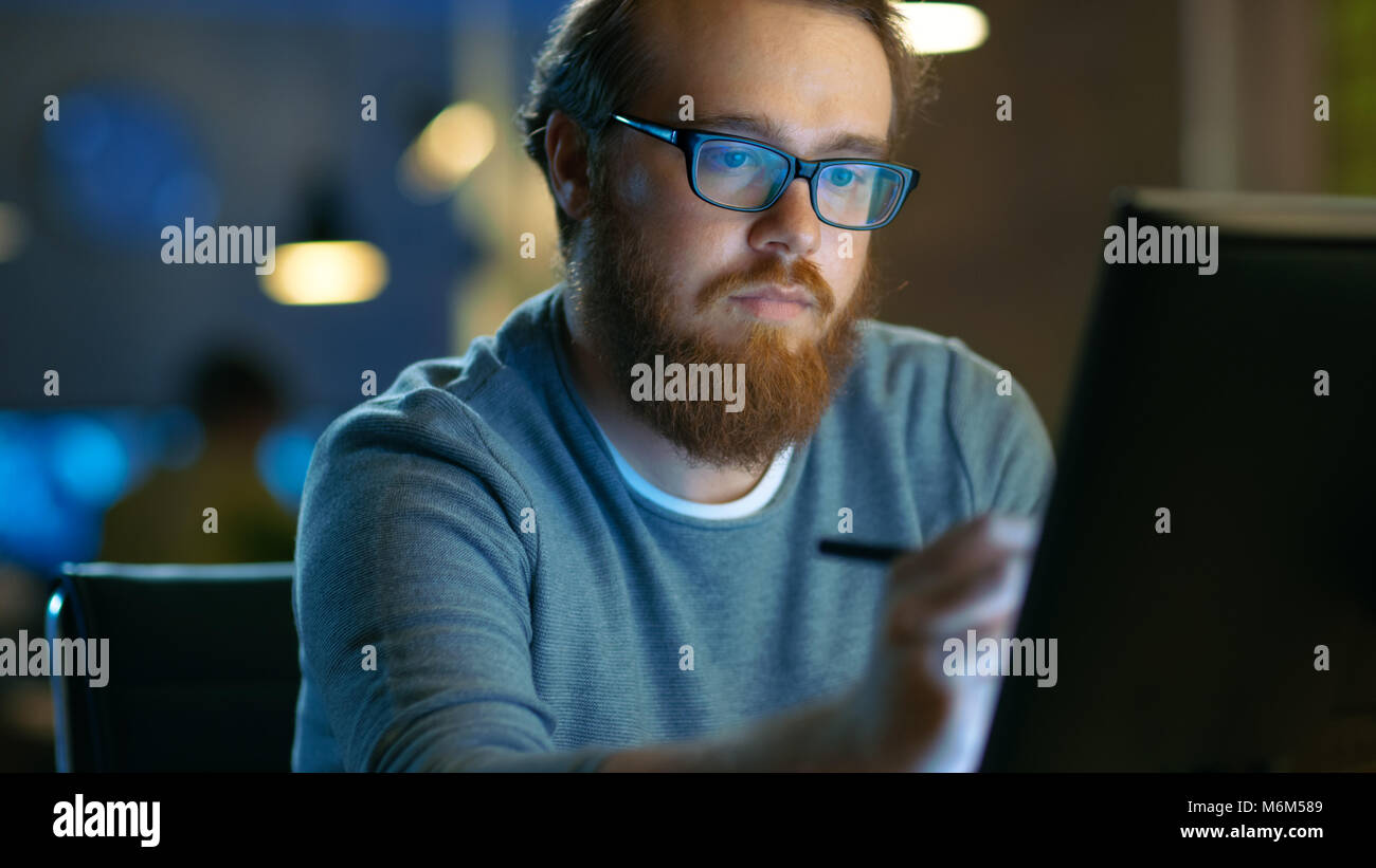 Talented Young Concept Artist Drawing on a Digital Pen Display Tablet While Sitting at His Working Place in Modern Loft Office. Late at Night. Stock Photo