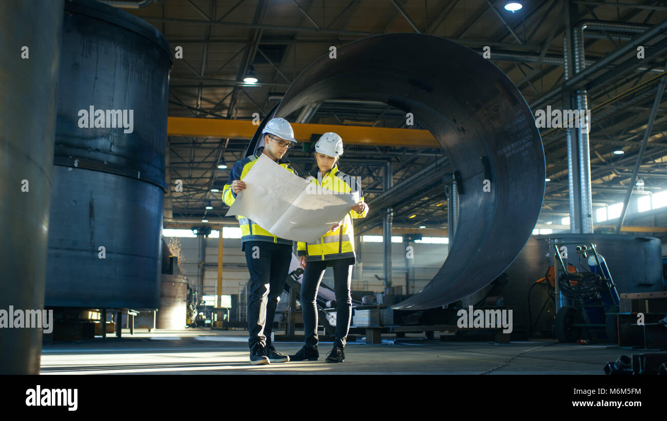 Male and Female Industrial Engineers Look at Project Blueprints While Standing Surround By Pipeline Parts in the Middle of Enormous Heavy Industry Stock Photo