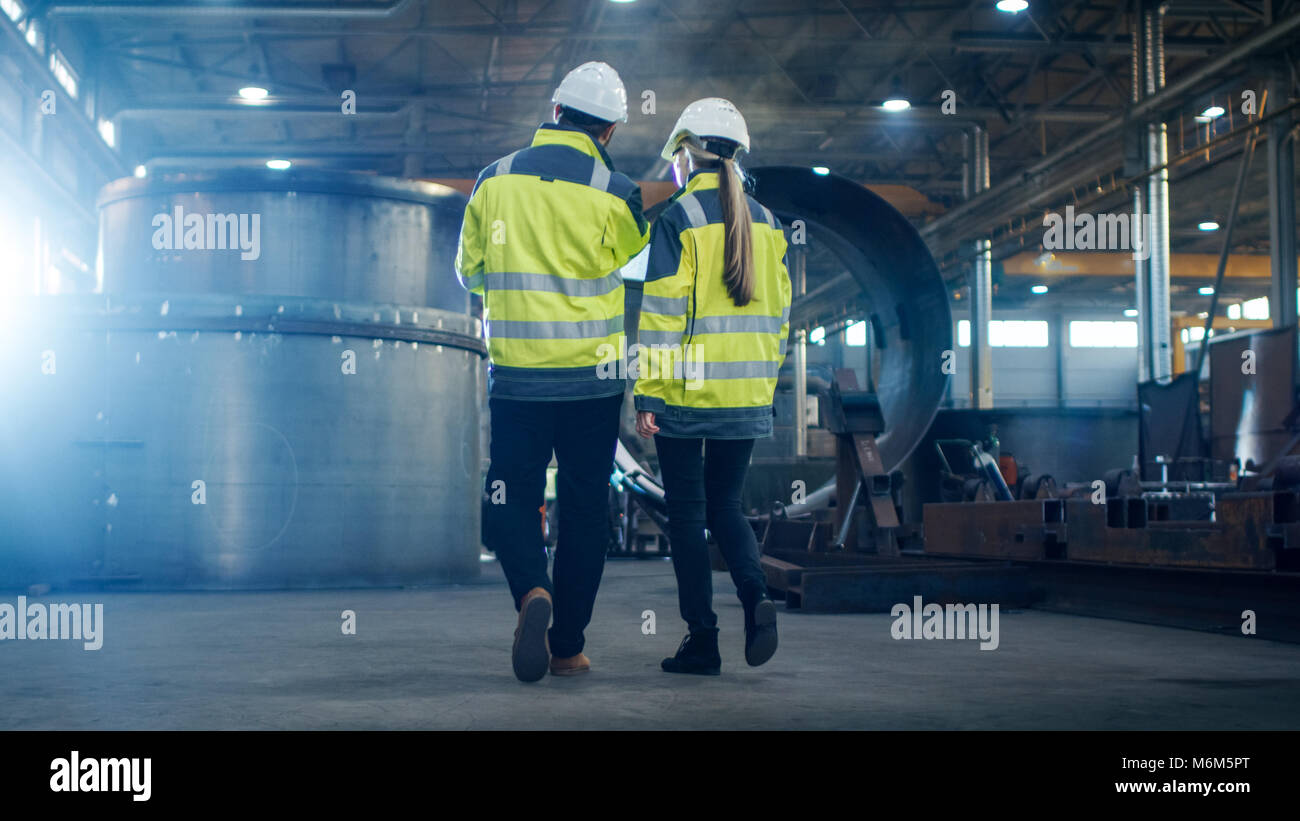 Male and Female Industrial Engineers use Laptop and Have Discussion While Walking Through Heavy Industry Manufacturing Factory. They Wear Hard Hats Stock Photo