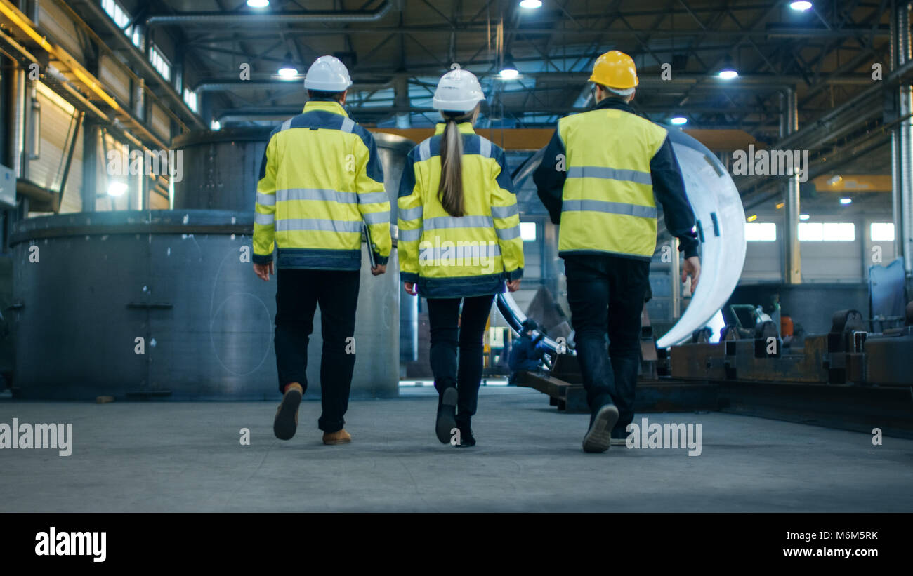 Following Shot of Three Engineers Walking Through Heavy Industry Manufacturing Factory. In the Background Welding Work in Progress, Various Metalwork Stock Photo