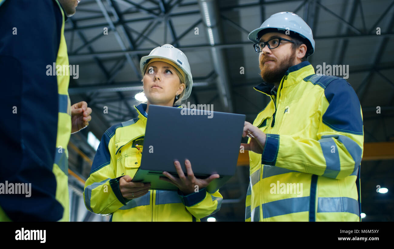 Male and Female Industrial Engineers Talk with Factory Worker while Using Laptop. They Work at the Heavy Industry Manufacturing Facility. Low Angle. Stock Photo