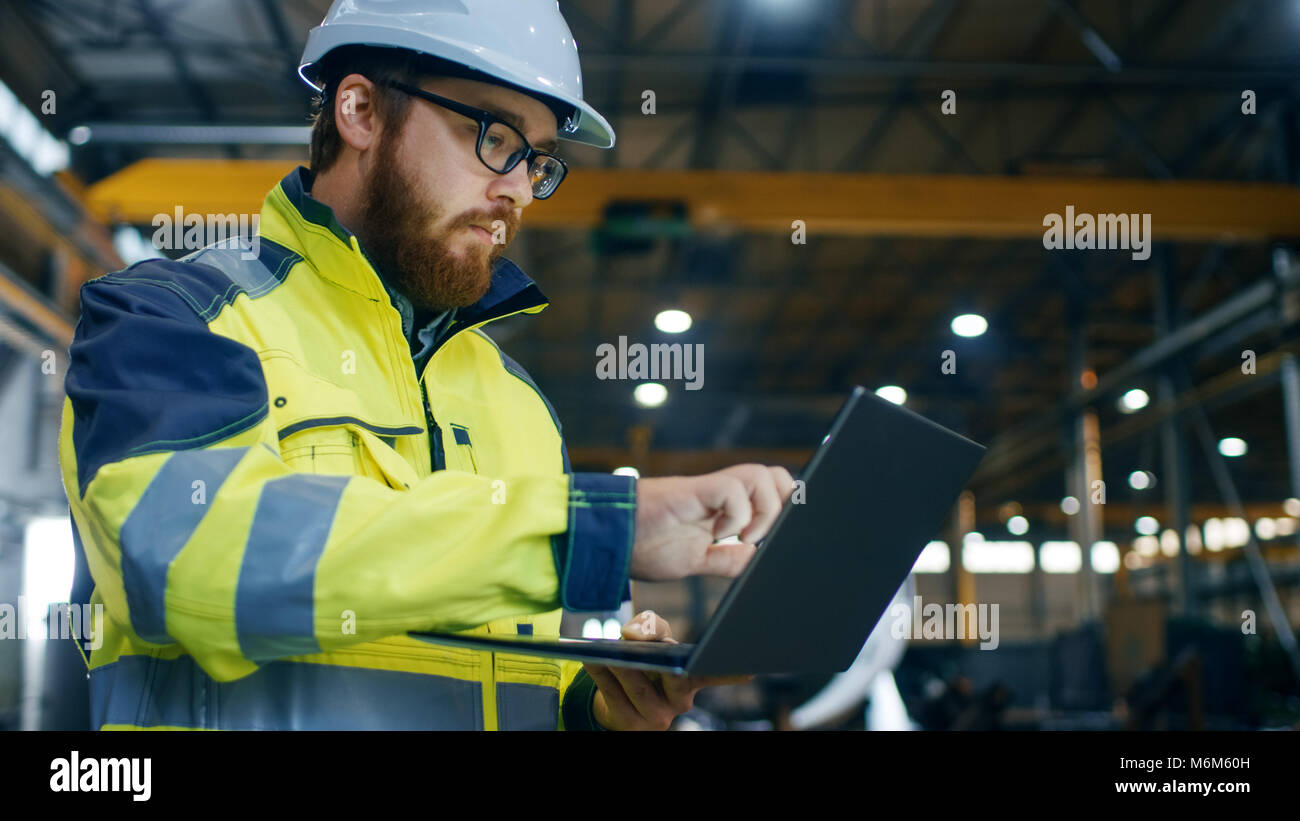 Industrial Engineer in Hard Hat Wearing Safety Jacket Uses Touchscreen Laptop. He Works at the Heavy Industry Manufacturing Factory. Stock Photo