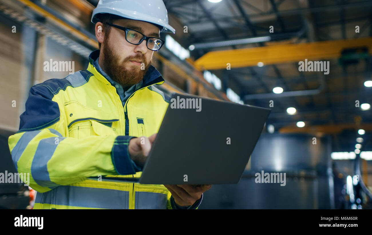 Industrial Engineer in Hard Hat Wearing Safety Jacket Uses Touchscreen Laptop. He Works at the Heavy Industry Manufacturing Factory. Stock Photo