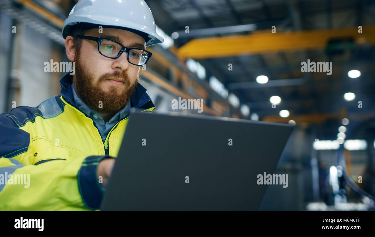 Industrial Engineer in Hard Hat Wearing Safety Jacket Uses Touchscreen Laptop. He Works at the Heavy Industry Manufacturing Factory. Stock Photo