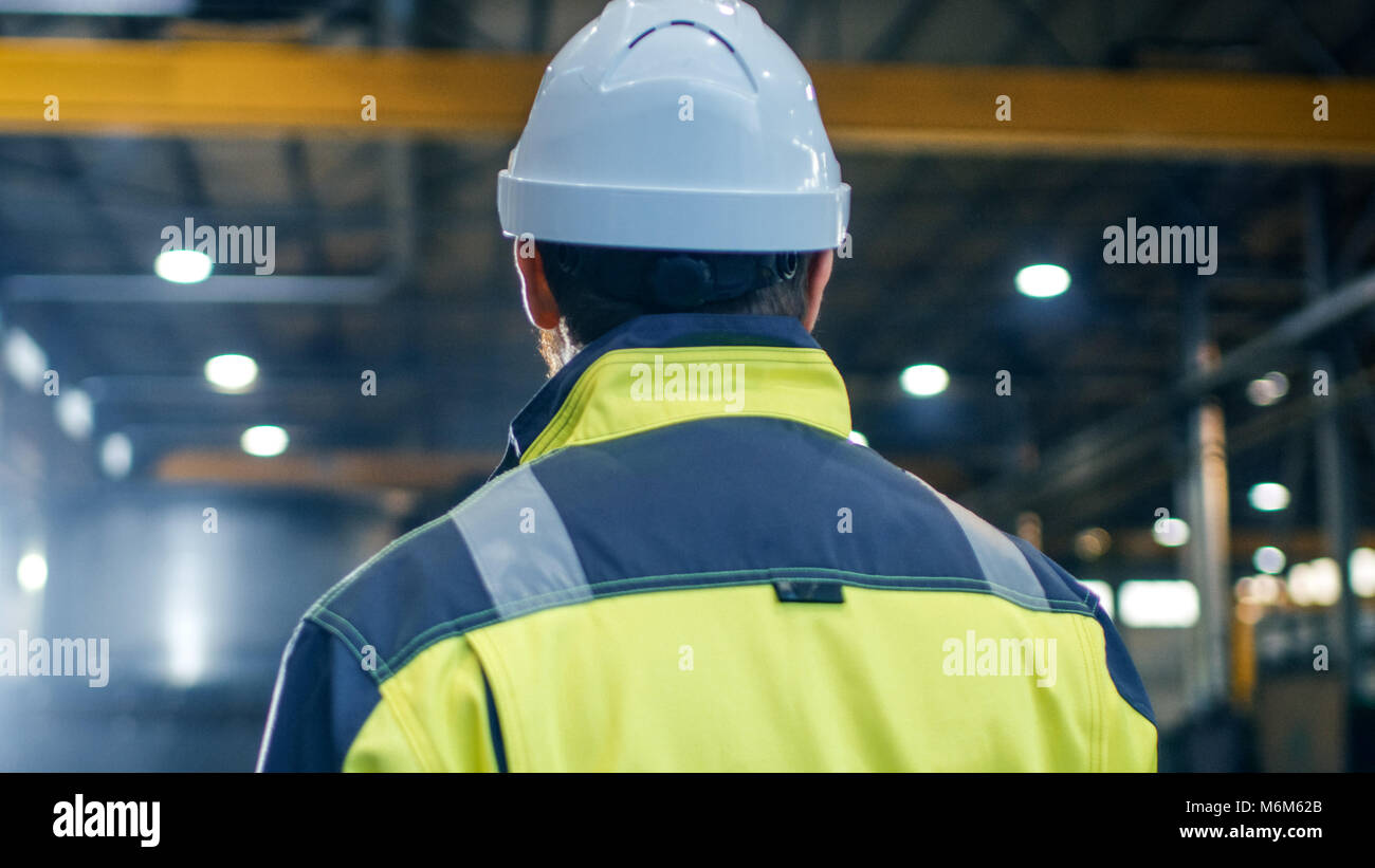 Industrial Engineer in Hard Hat and Safety Jacket Looks Around in Big Heavy Industry Manufacturing Factory. Stock Photo