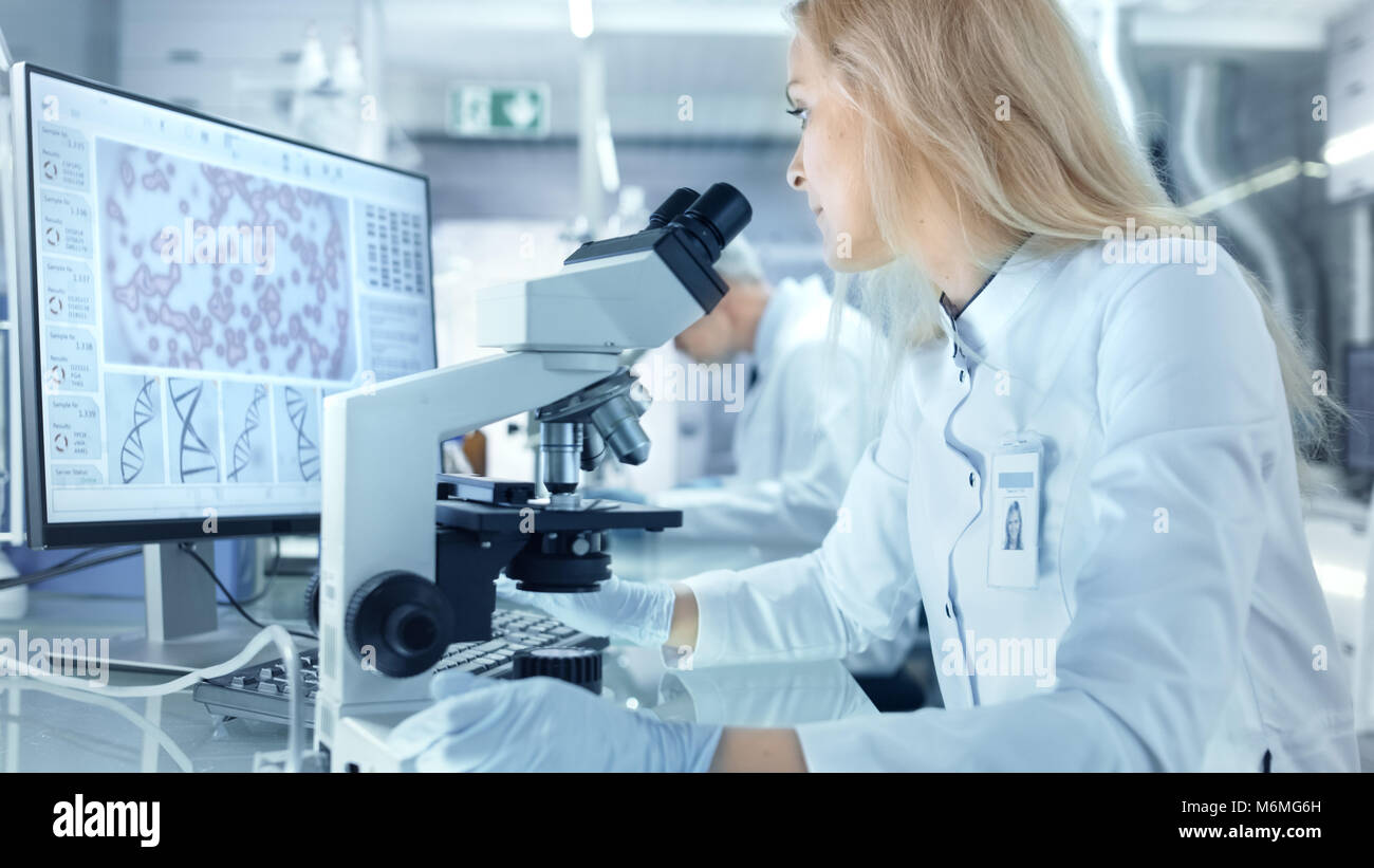 Female Research Scientist Using Electronic Microscope. She and Her Colleagues Work in a Big Modern Laboratory/ Medical Centre. Stock Photo