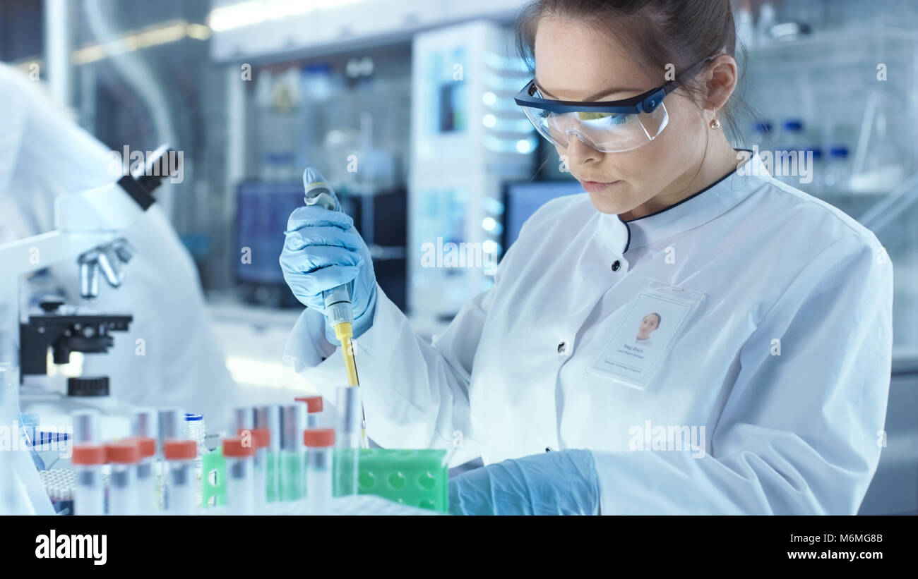 Female Research Scientist Uses Micropipette Filling Test Tubes in a Big Modern Laboratory. In the Background Scientists are Working. Stock Photo