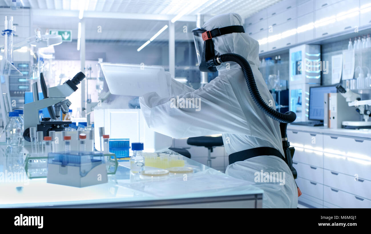Medical Virology Research Scientist Works in a Hazmat Suit with Mask, She Takes out Test  Tubes from Refrigerator Box. She Works in a Sterile Lab Stock Photo